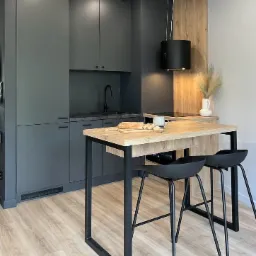Nowoczesna kuchnia w odcieniach szarości z laminowanymi frontami, drewnianym blatem i czarnymi akcentami, w tym minimalistycznym okapem i wysokimi krzesłami barowymi.