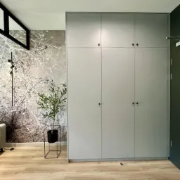 Szara szafa z płyty laminowanej, z sześcioma drzwiami i okrągłymi, czarnymi uchwytami, na tle ściany z tapetą w motywy drzewne, z widoczną rośliną doniczkową w czarnej, metalowej osłonce.