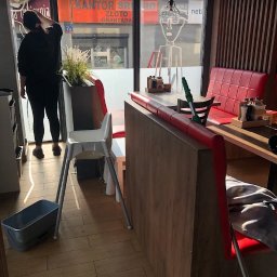 Czyste24 - Sprzątanie lokalu gastronomicznego: kobieta myje okno, widoczne krzesło dla dziecka, mop i wiadro. Lokal z czerwonymi kanapami i drewnianymi elementami.
