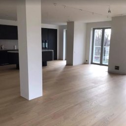 Czyste24 - Jasne, drewniane podłogi w przestronnym, minimalistycznym wnętrzu z czarną zabudową kuchenną i dużym oknem balkonowym. Widok na zewnątrz.