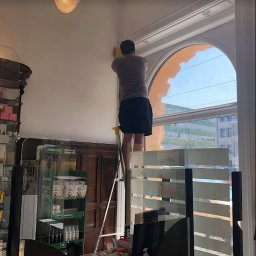 Czyste24 - Mężczyzna na drabinie czyści ramę okienną w pomieszczeniu z przeszklonymi przegrodami. Widok na budynki za oknem. Żółta ściereczka w dłoni.