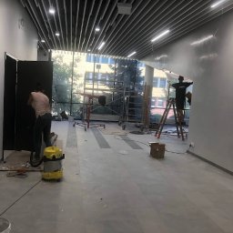 Czyste24 - Wnętrze biura w trakcie prac wykończeniowych: malowanie ścian, montaż paneli. Widoczne rusztowania, drabina i odkurzacz przemysłowy. Duże okna z widokiem na budynek.