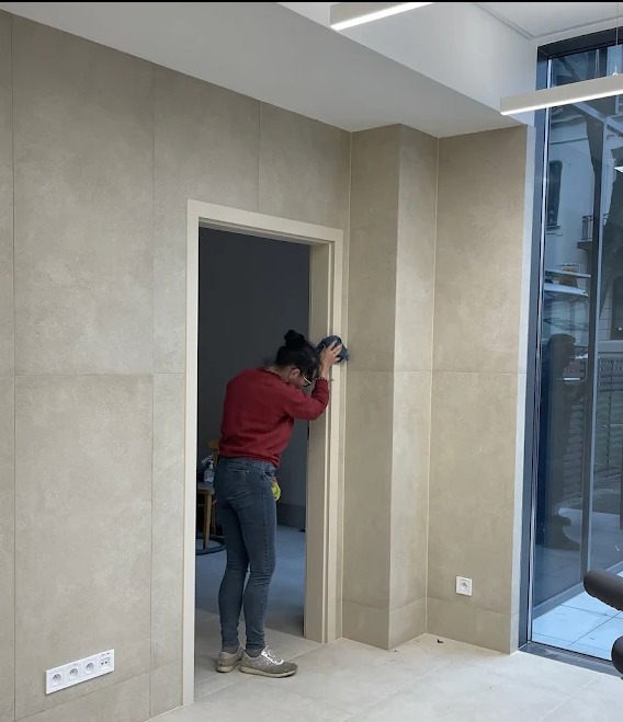 Kobieta w czerwonej bluzie czyści framugę drzwi w jasnym wnętrzu. Widoczne płytki na ścianach i podłodze, gniazdka elektryczne. Praca wykonywana z użyciem niebieskiej ściereczki.