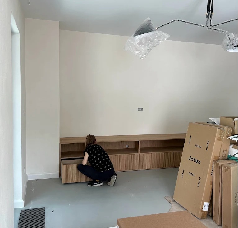 Montaż mebli: kobieta instaluje szafkę RTV z lamelami w nowym wnętrzu. Kartony JOTEX obok. Lampa sufitowa w folii ochronnej. Szare podłoże.