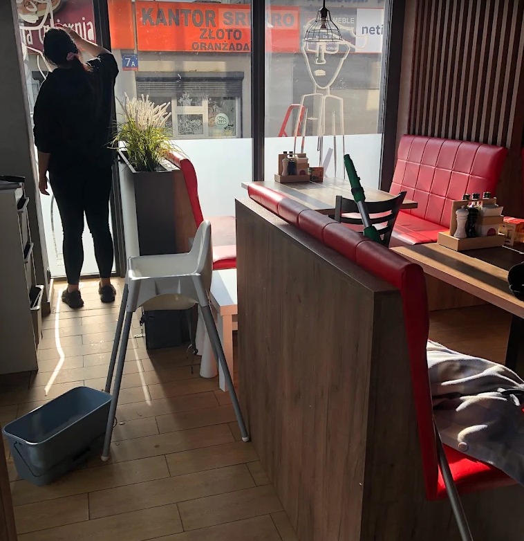 Sprzątanie lokalu gastronomicznego: kobieta myje okno, widoczne krzesło dla dziecka, mop i wiadro. Lokal z czerwonymi kanapami i drewnianymi elementami.