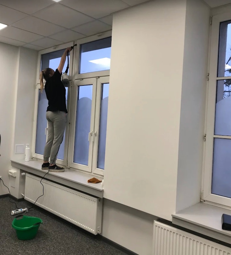 Kobieta w szarych spodniach i czarnej koszulce myje okno za pomocą specjalistycznego sprzętu. Widoczne wiadro i inne akcesoria na parapecie.