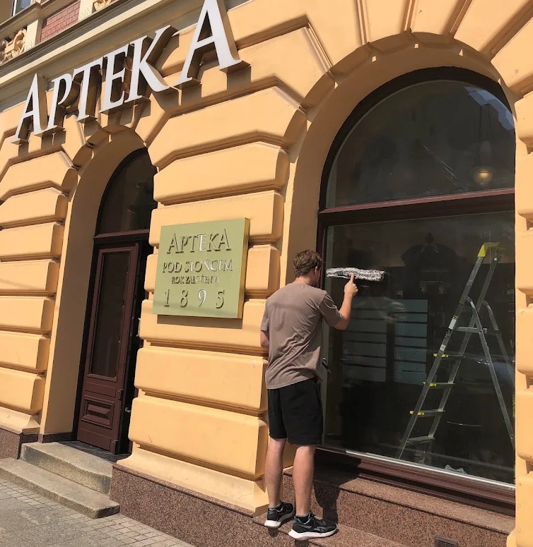 Mężczyzna myje okno apteki z użyciem myjki. Widoczna drabina oparta o ścianę budynku. Na fasadzie szyld z nazwą apteki i rokiem założenia 1895.