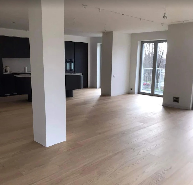 Jasne, drewniane podłogi w przestronnym, minimalistycznym wnętrzu z czarną zabudową kuchenną i dużym oknem balkonowym. Widok na zewnątrz.