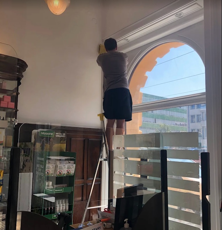 Mężczyzna na drabinie czyści ramę okienną w pomieszczeniu z przeszklonymi przegrodami. Widok na budynki za oknem. Żółta ściereczka w dłoni.