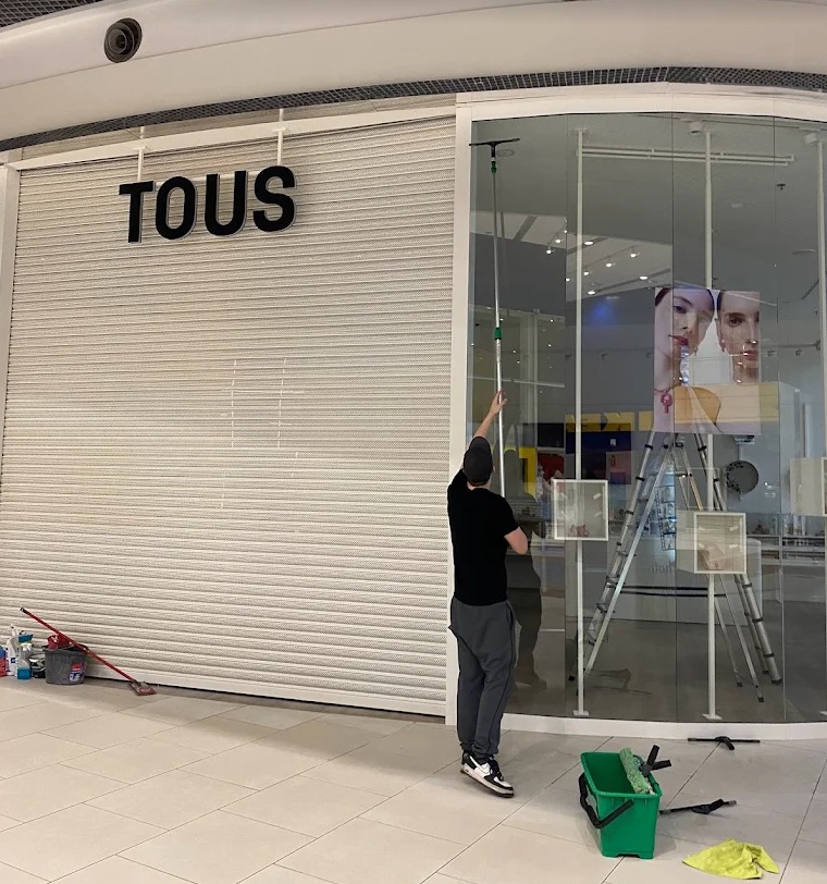 Pracownik myje witrynę sklepu TOUS w galerii handlowej. Widoczny sprzęt do mycia okien, drabina i zamknięta roleta. Ujęcie z dołu, perspektywa z poziomu podłogi.