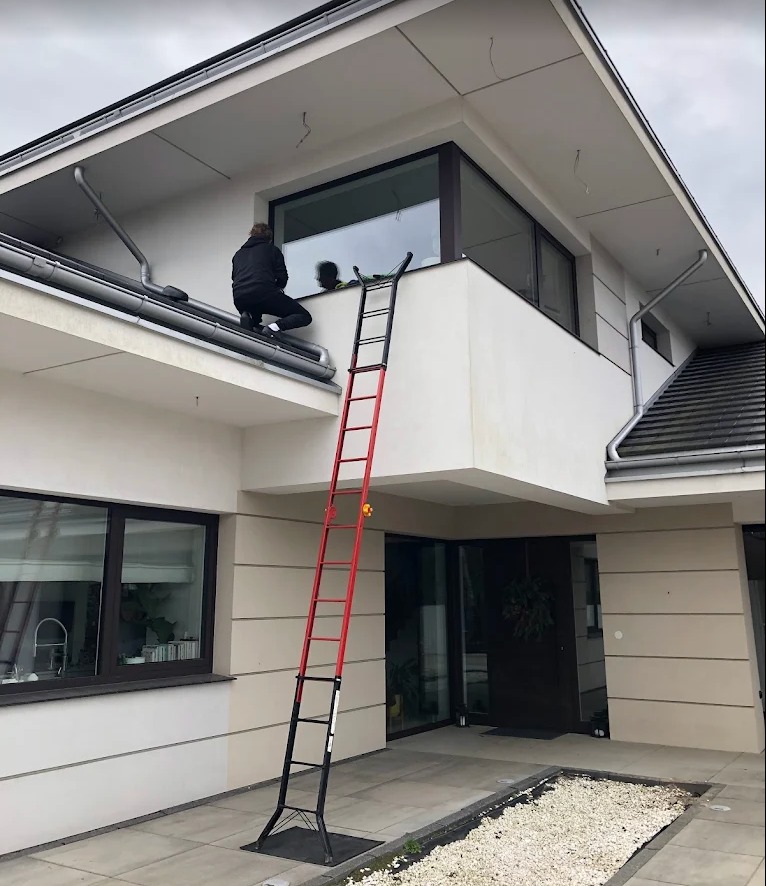 Mycie okien na wysokości w nowoczesnym domu. Pracownik siedzi na dachu, drugi na drabinie. Widoczne okno z odbiciem i elewacja budynku.