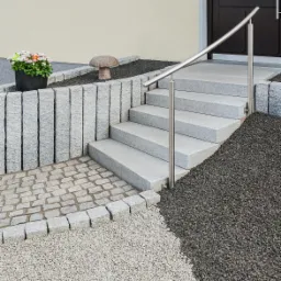 Zewnętrzne schody kamienne z nierdzewną balustradą prowadzące do drzwi wejściowych, otoczone różnymi rodzajami kamieni i roślinnością, w tym doniczką z kwiatami i kamiennym grzybkiem.