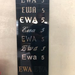 P.H.U. ANTKOWIAK BARBARA - Czarna tablica z wygrawerowanymi napisami 'EWA 5' w różnych stylach czcionek i kolorach, odbicie fotografa widoczne na powierzchni.