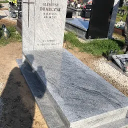 Jasnoszary granitowy nagrobek z wygrawerowanym krzyżem i inskrypcją na cmentarzu, widok z boku, otoczony innymi nagrobkami.