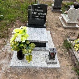 MARSTONE ANTKOWIAK BARBARA - Granitowy nagrobek z czarną tablicą i jasnoszarą podstawą, ozdobiony kwiatami w wazonie i lampionem, na cmentarzu z innymi nagrobkami w tle.