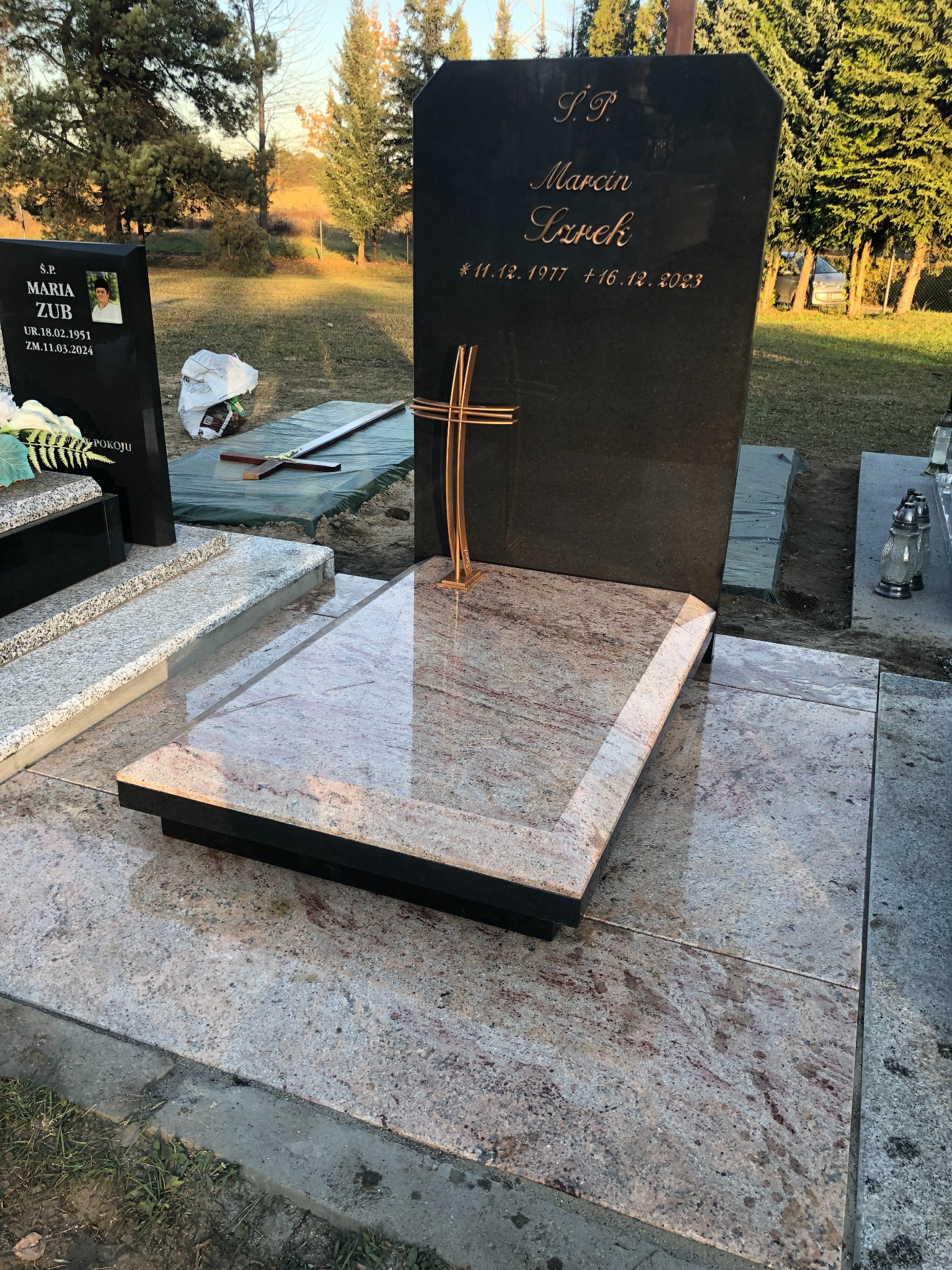 Nowy, ciemny nagrobek z inskrypcjami i złotym krzyżem, ustawiony na płycie z jasnego, wzorzystego kamienia, na tle cmentarza z drzewami.