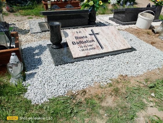 Granitowy nagrobek z jasną płytą inskrypcyjną, otoczony białym grysem, na cmentarzu w słoneczny dzień.