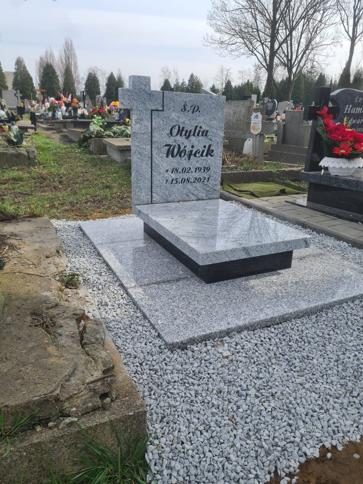 Granitowy pomnik z krzyżem i napisem 'Ś.P. Otylia Wójcik' oraz datami urodzin i śmierci, otoczony szarym żwirem na cmentarzu.
