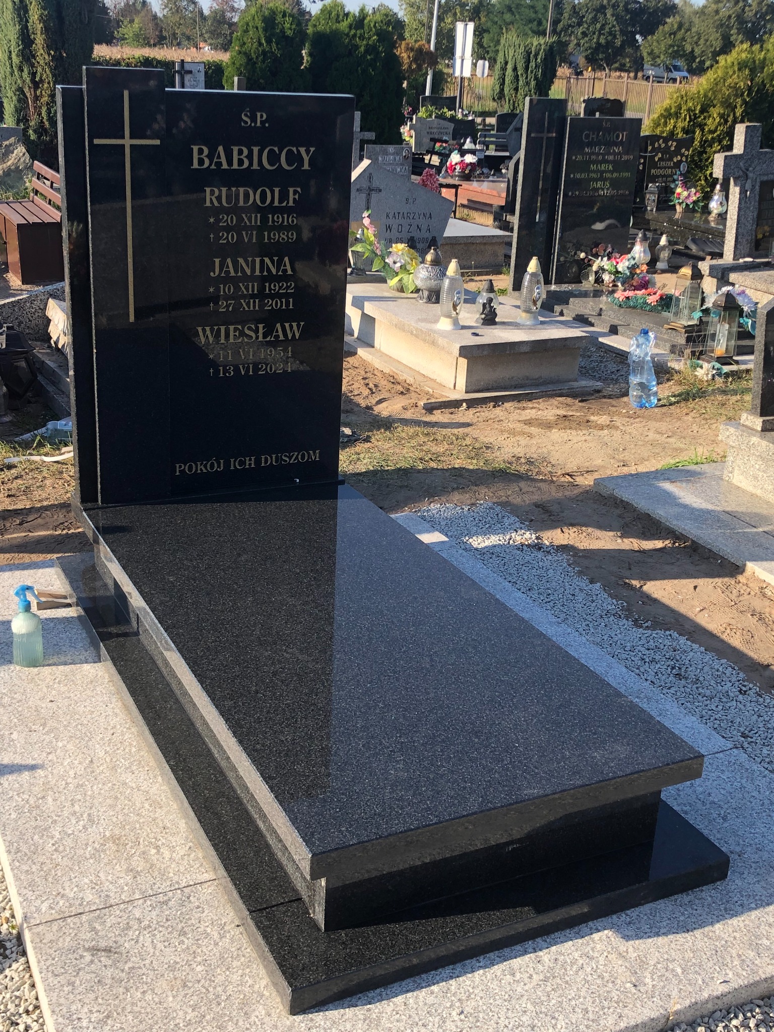 Czarny, granitowy pomnik nagrobny z wygrawerowanymi złotymi literami i krzyżem, ustawiony na cmentarzu, obok butelka z płynem do czyszczenia.