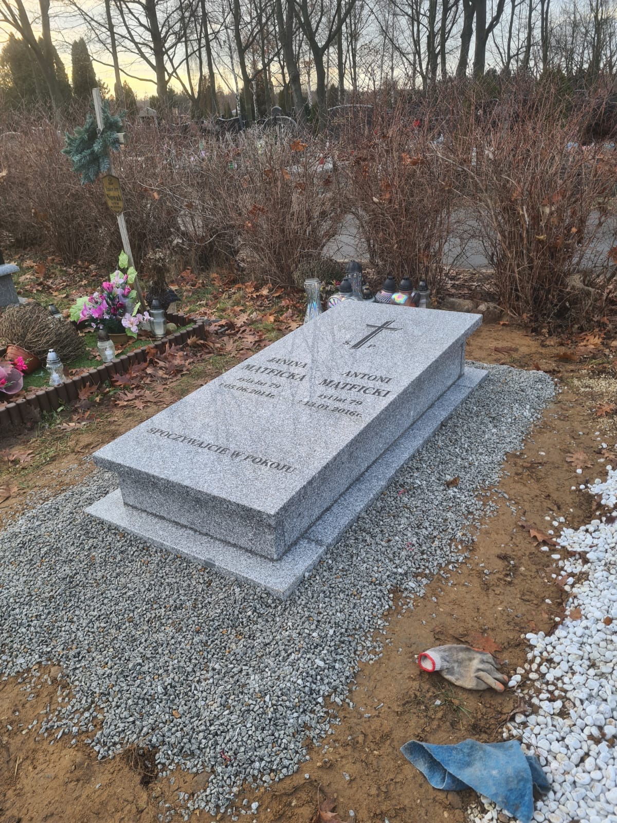 Podwójny, jasnoszary pomnik z wygrawerowanym krzyżem i inskrypcjami na cmentarzu, otoczony drobnym żwirem, z rękawicą roboczą i szmatką leżącą obok.