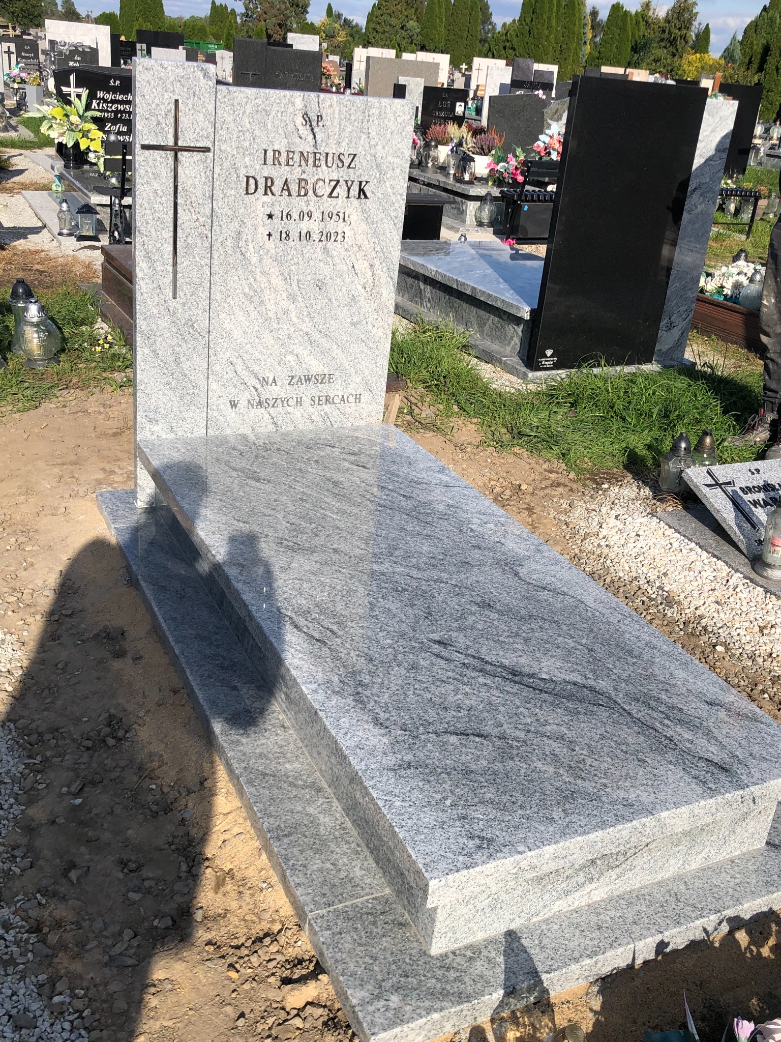 Jasnoszary granitowy nagrobek z wygrawerowanym krzyżem i inskrypcją na cmentarzu, widok z boku, otoczony innymi nagrobkami.