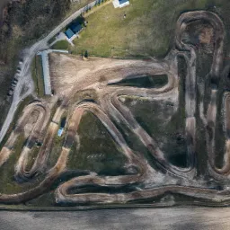 Widok z drona na tor motocrossowy z licznymi zakrętami i śladami opon, otoczony polami i zabudowaniami gospodarczymi, w słoneczny dzień.