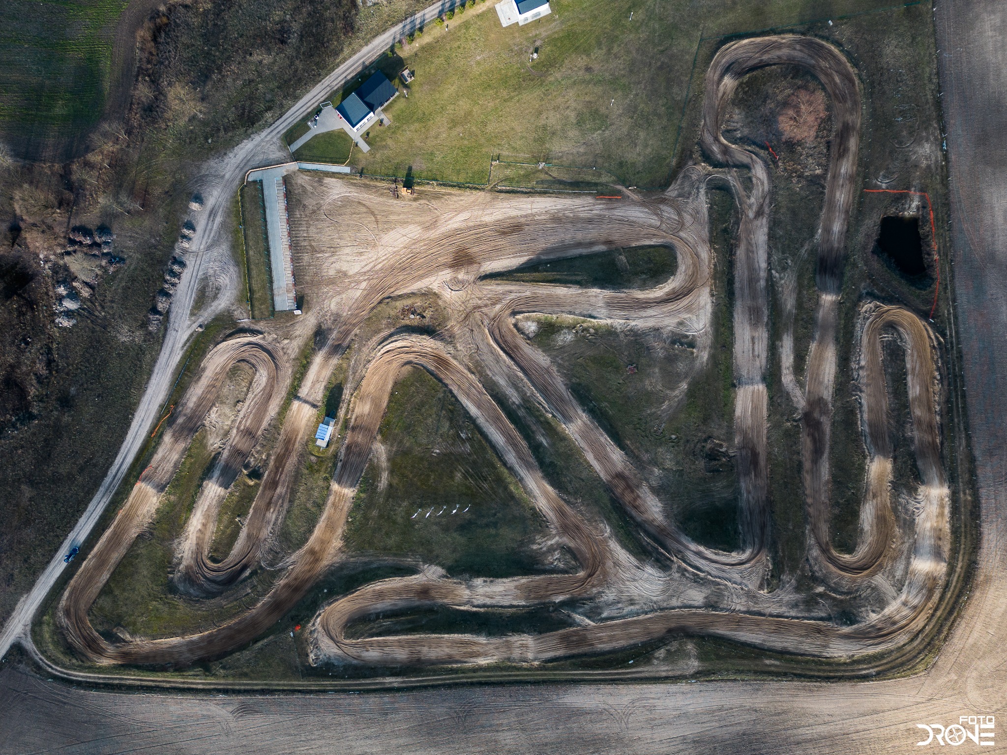 Widok z drona na tor motocrossowy z licznymi zakrętami i śladami opon, otoczony polami i zabudowaniami gospodarczymi, w słoneczny dzień.