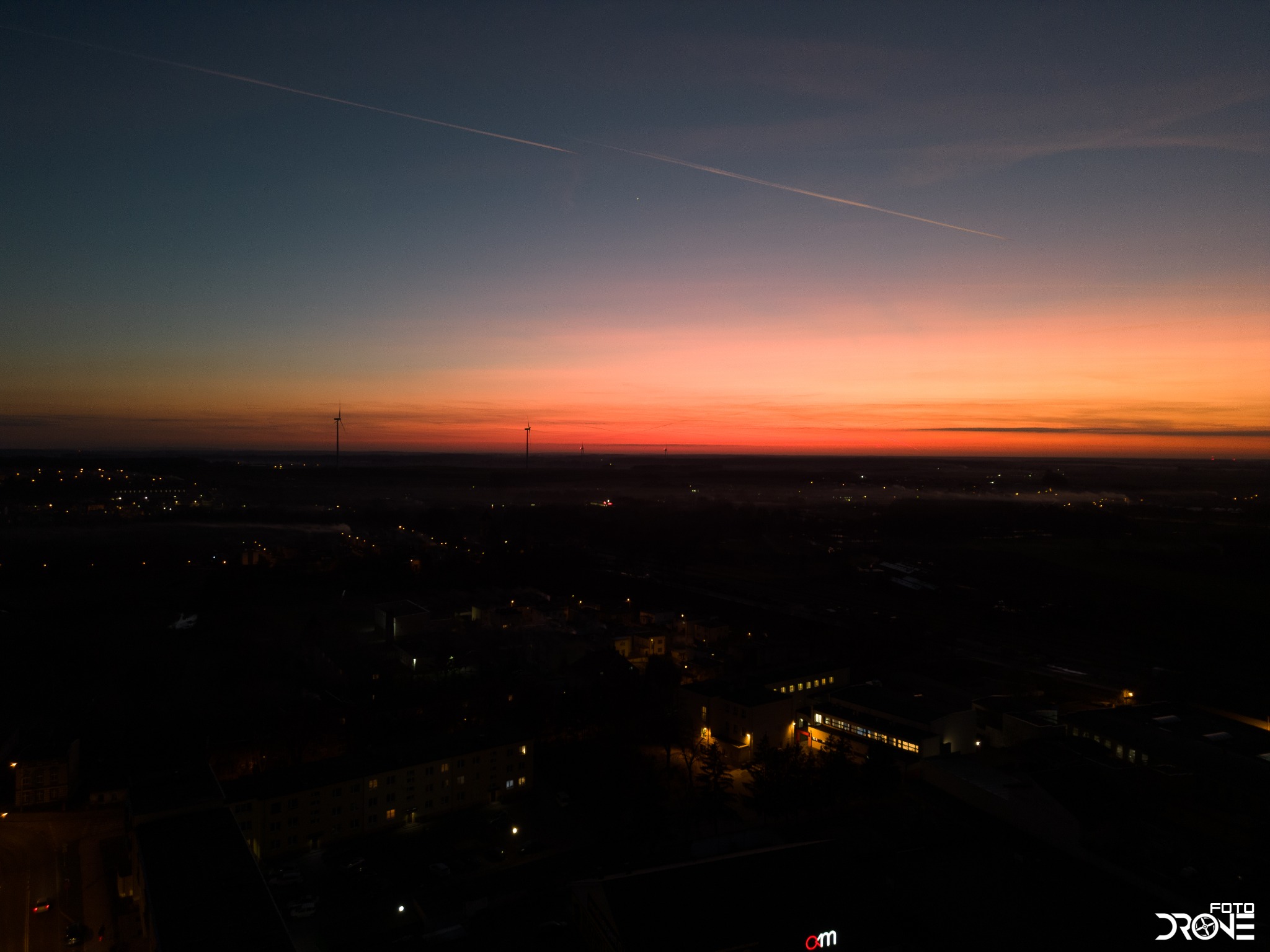 Nocny widok z drona na miasto oświetlone słabymi lampami, z pomarańczowo-różowym niebem w tle i smugą kondensacyjną samolotu.