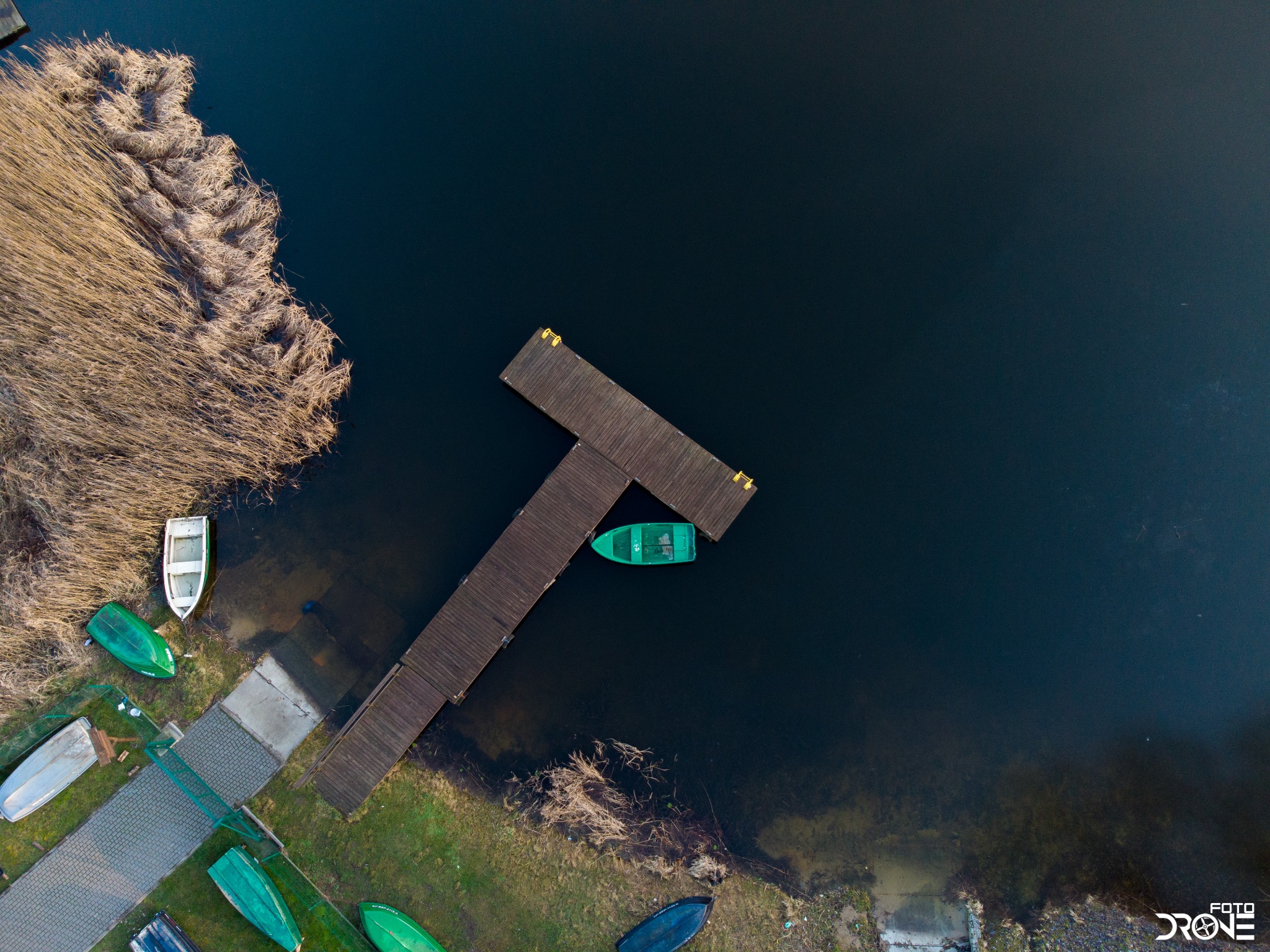 Widok z drona na pomost w kształcie litery T, zieloną łódkę zacumowaną przy pomoście, oraz kilka łódek na brzegu jeziora otoczonego trzcinami i trawą.