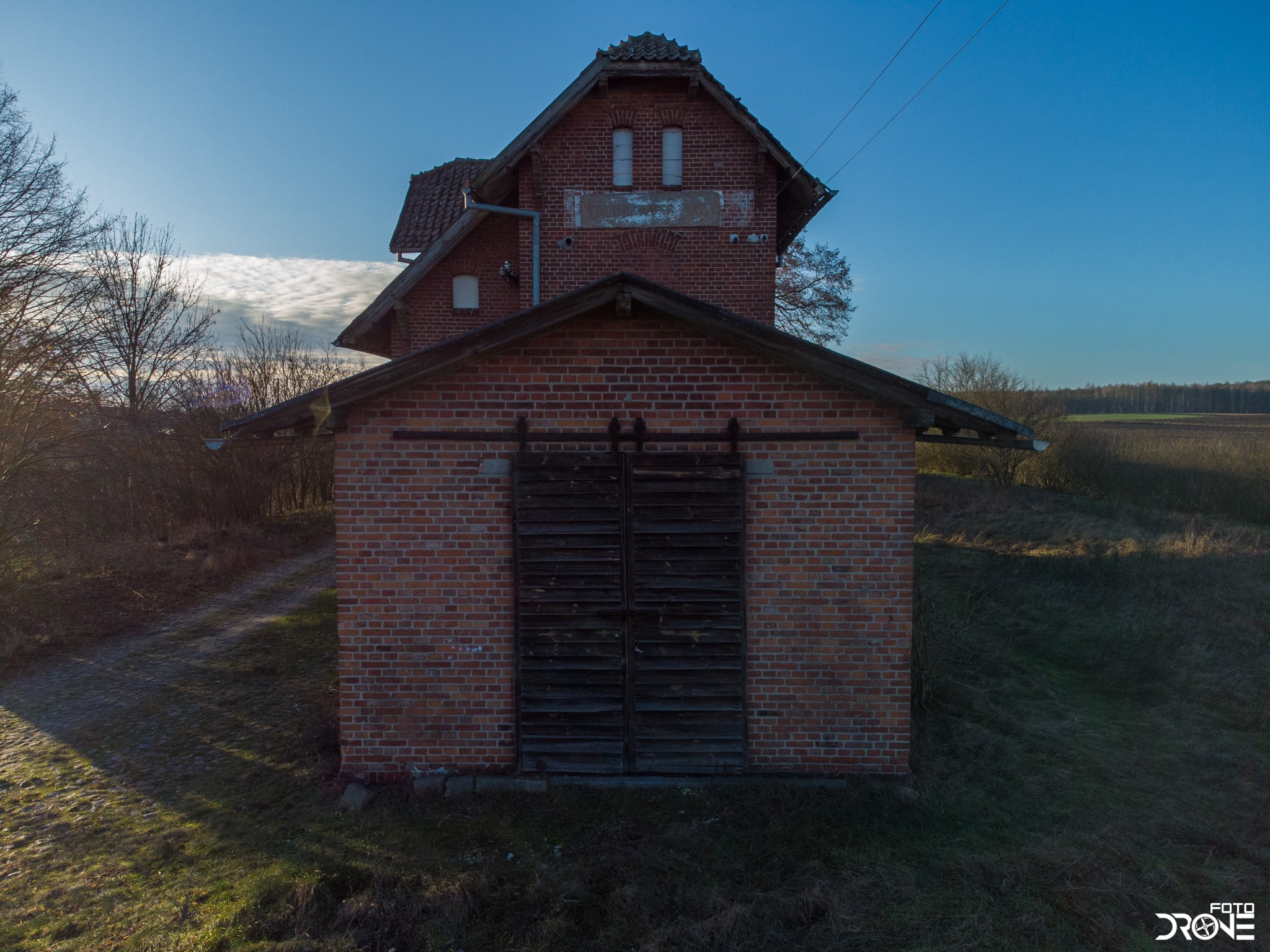 Dwukondygnacyjny, opuszczony budynek z czerwonej cegły z zarośniętym otoczeniem, sfotografowany w słoneczny dzień.