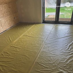 HUAR spółka z ograniczoną odpowiedzialnością - Wnętrze domu w trakcie budowy z widoczną izolacją podłogową w kolorze żółtym, zabezpieczającą przed wilgocią, ściany wykończone płytą OSB i gładzią, okno balkonowe z widokiem na zieleń.