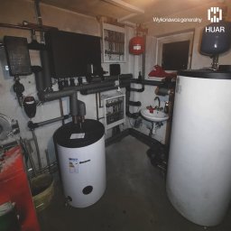HUAR spółka z ograniczoną odpowiedzialnością - Kotłownia z dwoma białymi zbiornikami na wodę, licznymi rurami, zaworami i małą umywalką w tle, oświetlona sztucznym światłem.