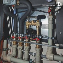 HUAR spółka z ograniczoną odpowiedzialnością - Węzeł instalacji grzewczej z licznymi zaworami, rurami miedzianymi i plastikowymi, oraz czarnymi przewodami w kotłowni w Warszawie.