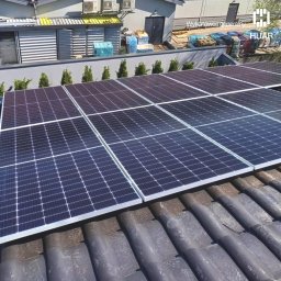 HUAR spółka z ograniczoną odpowiedzialnością - Panele słoneczne na ciemnym, ceramicznym dachu, z widocznym budynkiem gospodarczym w tle i logiem firmy w rogu.