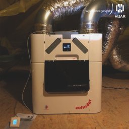 HUAR spółka z ograniczoną odpowiedzialnością - Centrala wentylacyjna Zehnder z podłączonymi przewodami wentylacyjnymi w pomieszczeniu z drewnianą podłogą, widoczny panel sterowania i logo firmy HUAR w prawym górnym rogu.