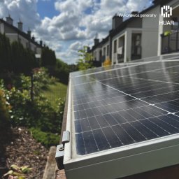 HUAR spółka z ograniczoną odpowiedzialnością - Panel fotowoltaiczny zamontowany na dachu domu jednorodzinnego, widok z bliska na mocowanie z użyciem śrub, w tle zabudowa szeregowa i fragment ogrodu.