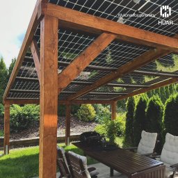 HUAR spółka z ograniczoną odpowiedzialnością - Drewniana pergola z panelami słonecznymi na dachu, widok z boku na stół i krzesła ogrodowe, w tle zadbany ogród z tujami i trawnikiem.