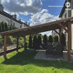 HUAR spółka z ograniczoną odpowiedzialnością - Drewniana pergola z panelami słonecznymi w ogrodzie, widok na domy w tle i błękitne niebo z chmurami.