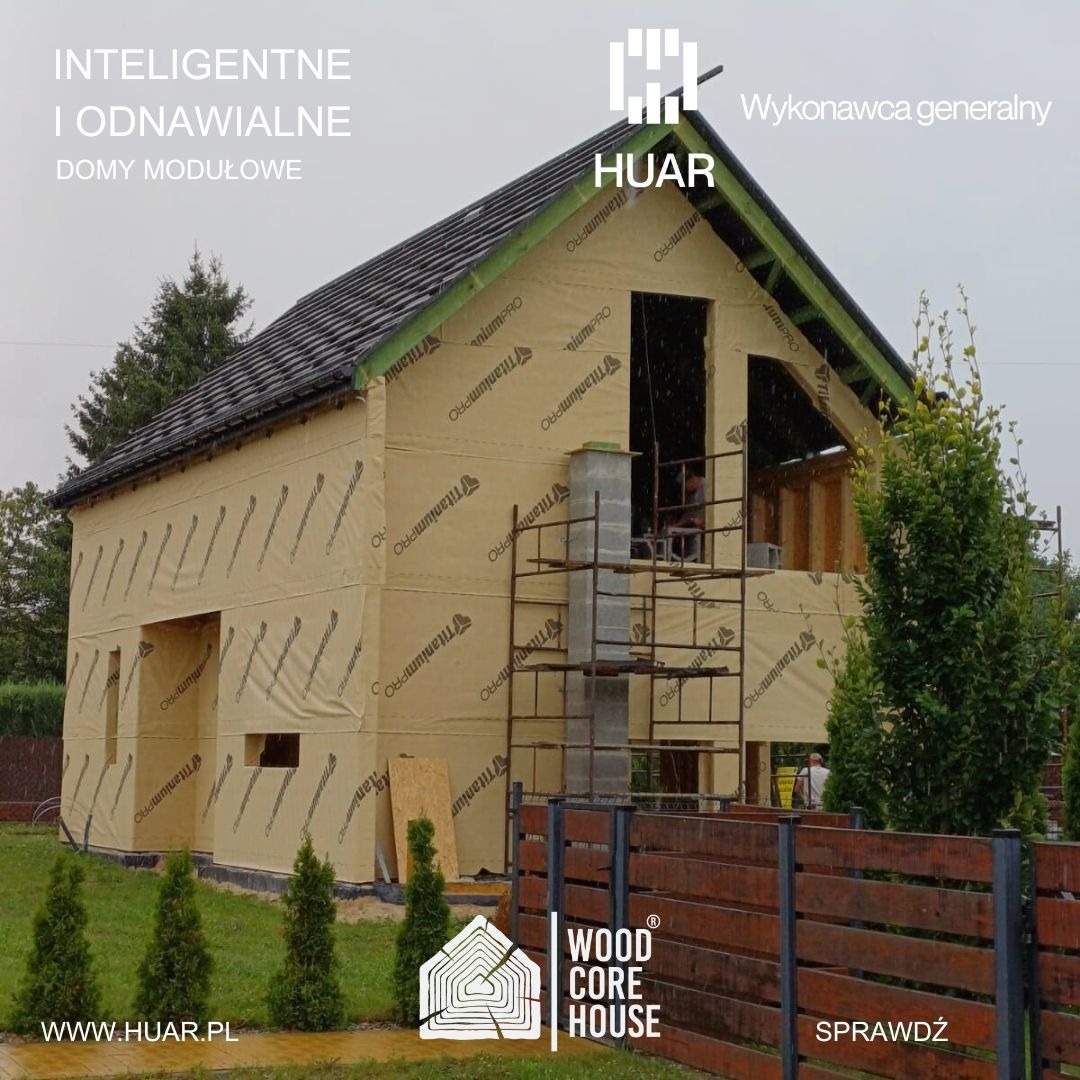 Budowa domu modułowego w trakcie wznoszenia, okrytego membraną izolacyjną z widocznym kominem i rusztowaniem, na pierwszym planie drewniane ogrodzenie i logo firmy Wood Core House.