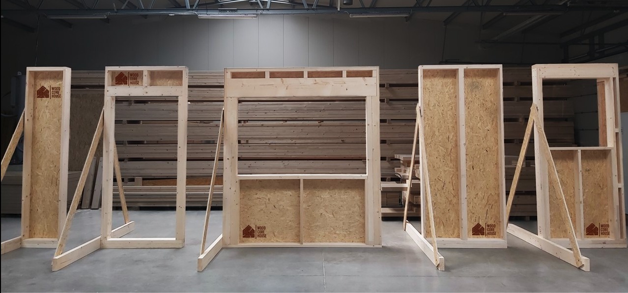 Szkieletowe elementy ścian z płyty OSB i drewna, ustawione w hali produkcyjnej, z widocznym logo producenta na każdej sekcji.