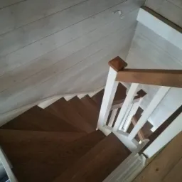 Wewnętrzne drewniane schody z białymi balustradami i brązowym poręczą, widok z góry na kręte stopnie, drewniana boazeria na ścianie i suficie w jasnym odcieniu.