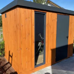 Drewniana budka z ciemną, płaską dachówką i ciemnymi drzwiami, z wąskim oknem, w którym widać rower w środku, otoczona zielenią i brukowanym chodnikiem.