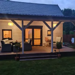 Oświetlony dom z białych paneli z zadaszonym tarasem i meblami ogrodowymi, widoczny z trawnika o zmierzchu.
