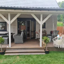 Zadaszony taras z drewna z meblami ogrodowymi z rattanu i huśtawką hamakową, na której siedzi uśmiechnięty chłopiec, widok na ogród z trawnikiem i roślinnością.