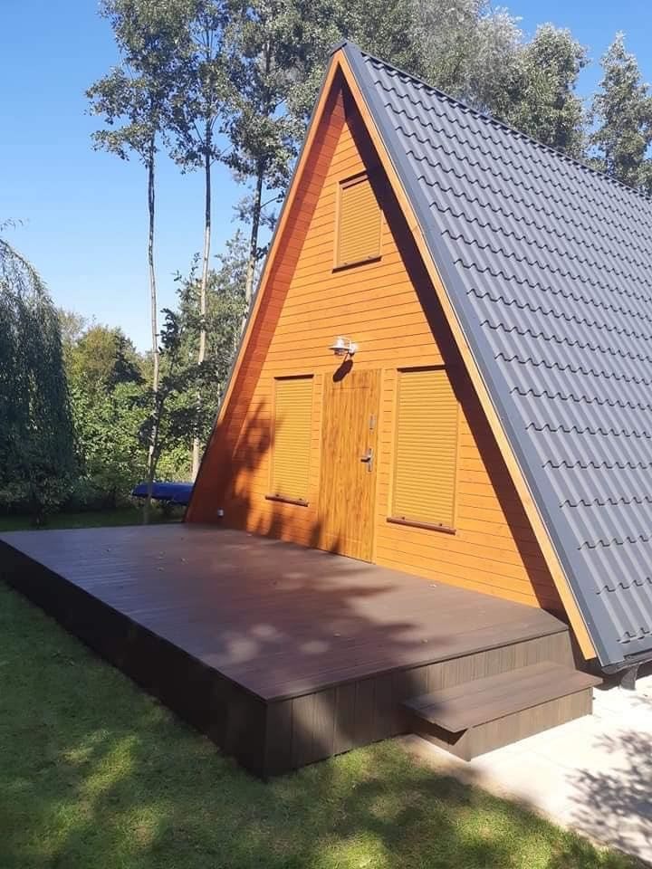 Nowoczesny, pomarańczowy domek letniskowy o trójkątnym kształcie z szarym dachem i dużym, brązowym tarasem kompozytowym, otoczony zielenią.