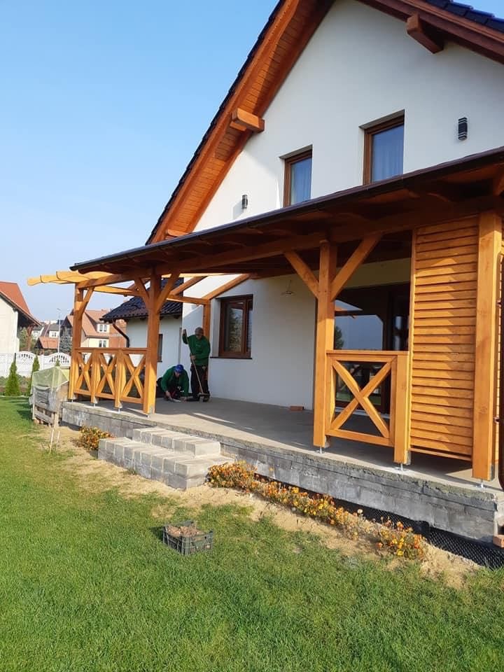 Drewniana pergola z balustradą przy białym domu jednorodzinnym; w tle pracownicy podczas montażu. Widoczne schody z kostki brukowej i rabata kwiatowa.