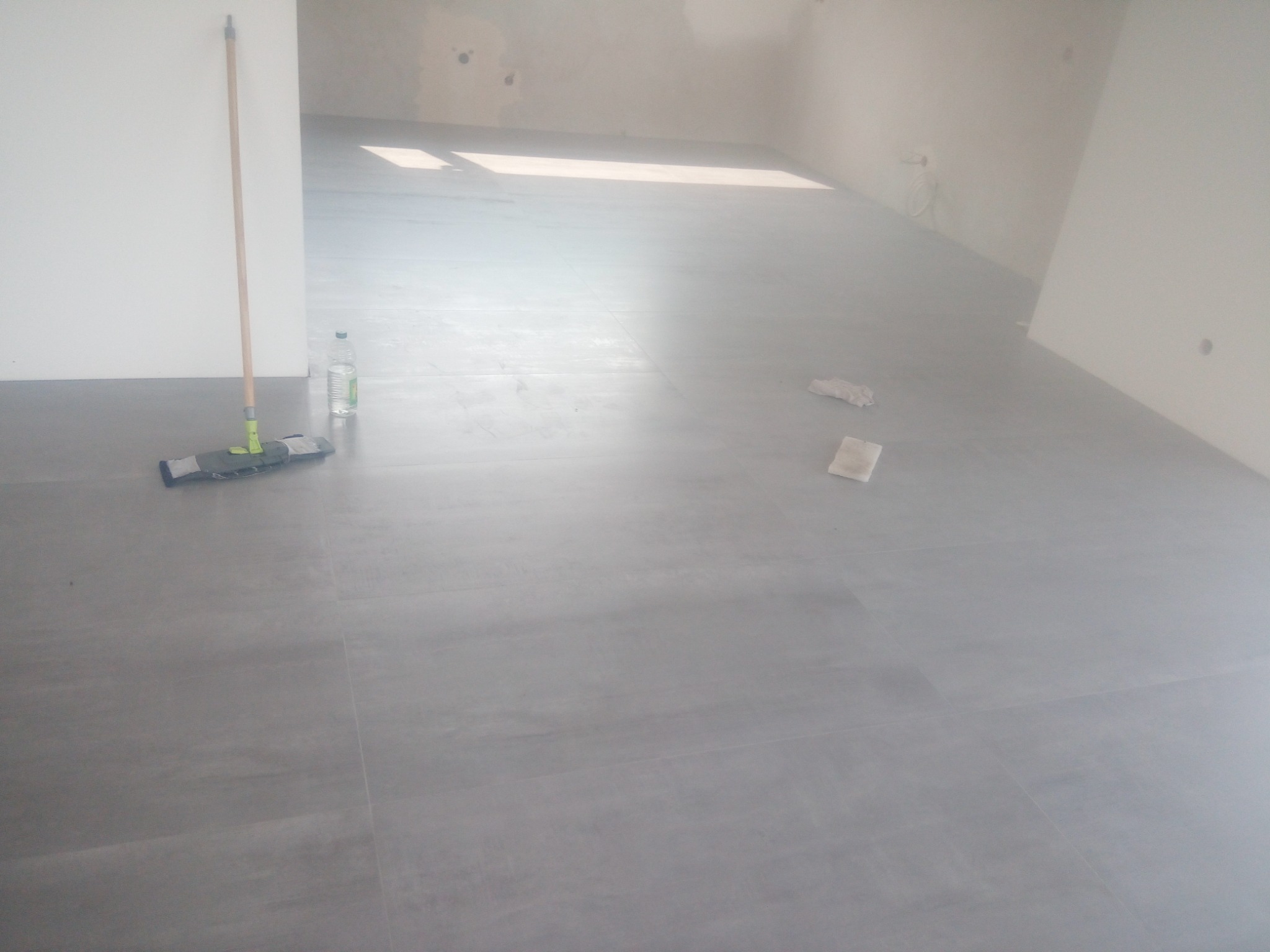 Szare płytki podłogowe w trakcie układania, widoczny mop i butelka z płynem do czyszczenia, ślady po zaprawie na ścianach.
