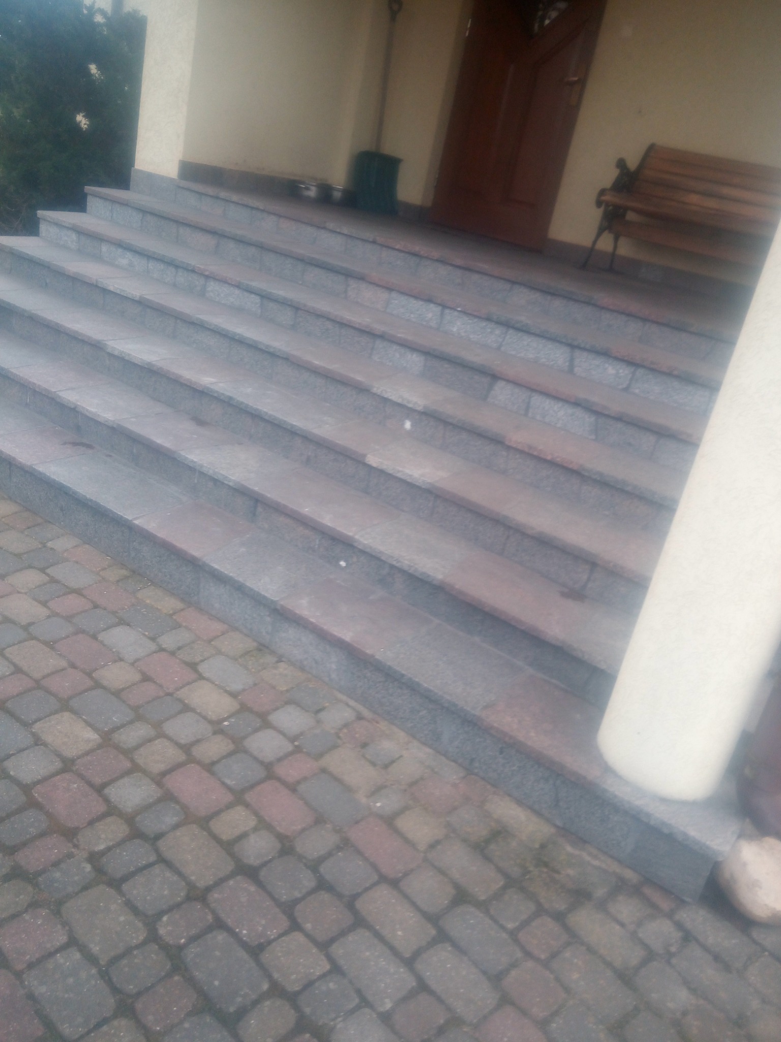 Zewnętrzne schody z szarego kamienia prowadzące do wejścia domu, z widoczną drewnianą ławką i kostką brukową na dole.