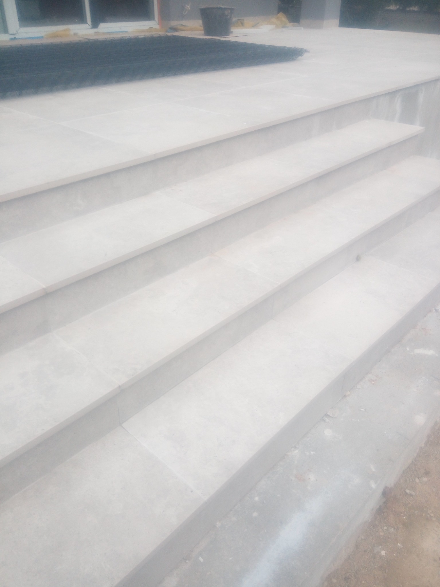 Zewnętrzne schody z jasnoszarych płytek, widoczne cztery stopnie, na górze mata ochronna i wiadro, niedokończone wykończenie po bokach.