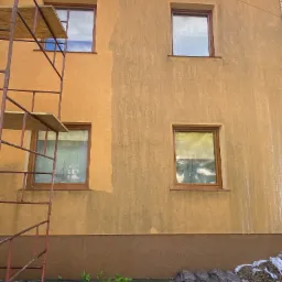 Fragment ściany budynku z oknami, częściowo umyta, z widocznym rusztowaniem i plamami brudu na elewacji, w tle błękitne niebo.